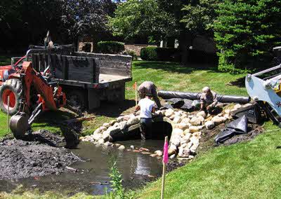 Workers arranging large rocks around a muddy pond in a residential yard